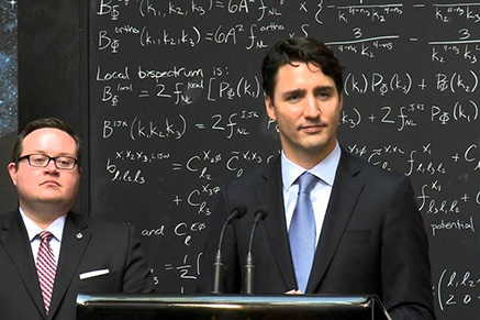 Trudeau stands in front of a background of equations on a chalk board